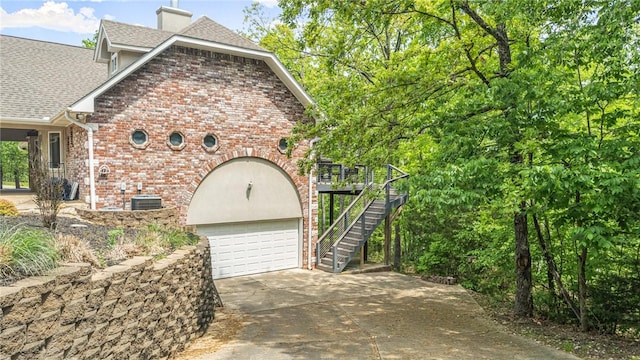 view of side of property with a shingled roof, brick siding, a garage, a chimney, and concrete driveway