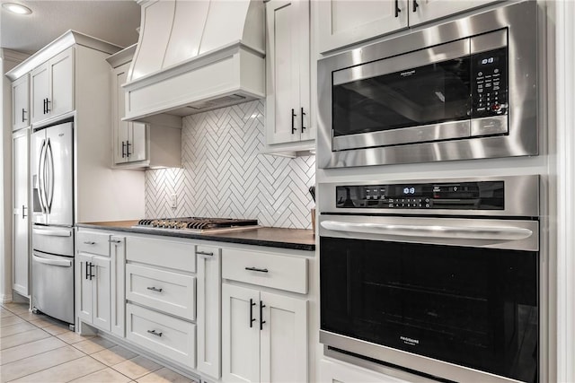 kitchen with custom exhaust hood, light tile patterned floors, dark countertops, backsplash, and appliances with stainless steel finishes