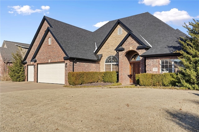 french country style house featuring a garage, brick siding, roof with shingles, and driveway