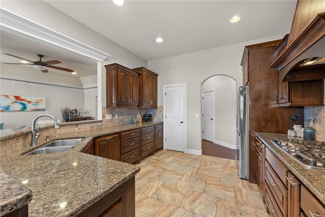 kitchen with arched walkways, custom exhaust hood, stainless steel appliances, a sink, and dark stone counters