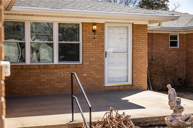 doorway to property featuring brick siding, roof with shingles, and a patio