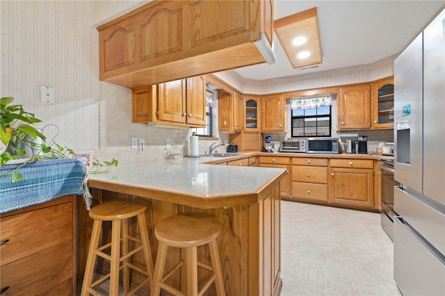 kitchen featuring wallpapered walls, glass insert cabinets, a breakfast bar, brown cabinets, and a peninsula