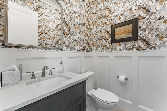 half bathroom with wallpapered walls, toilet, a decorative wall, a wainscoted wall, and vanity