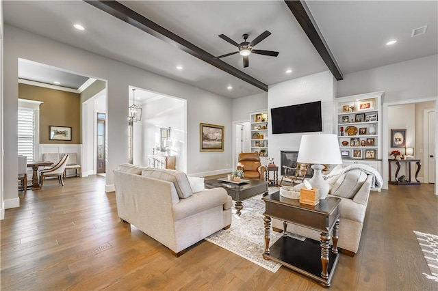 living room featuring beam ceiling, a ceiling fan, a glass covered fireplace, built in shelves, and wood finished floors
