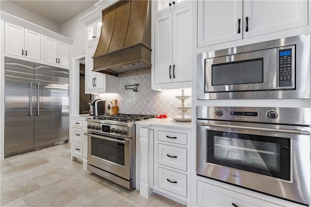 kitchen with built in appliances, custom range hood, light countertops, white cabinets, and backsplash