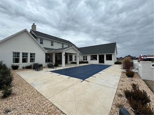 rear view of house with a covered pool, a chimney, roof with shingles, a patio area, and board and batten siding