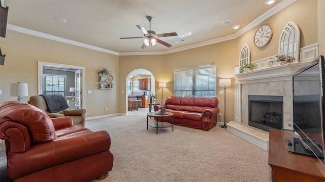 carpeted living area featuring arched walkways, ceiling fan, baseboards, a tiled fireplace, and crown molding