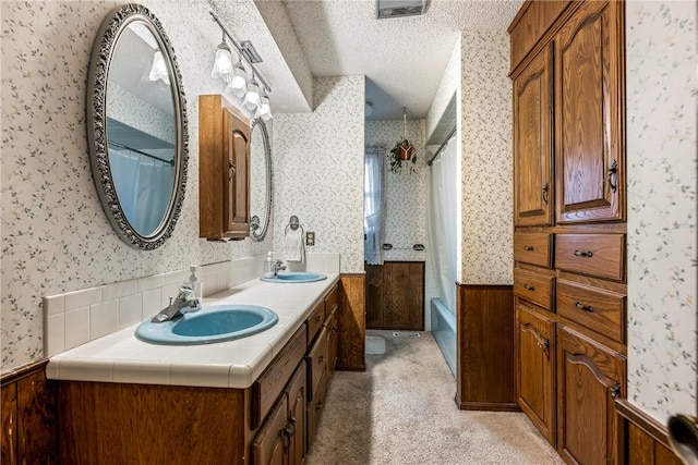 full bath with wallpapered walls, a textured ceiling, a sink, and wainscoting