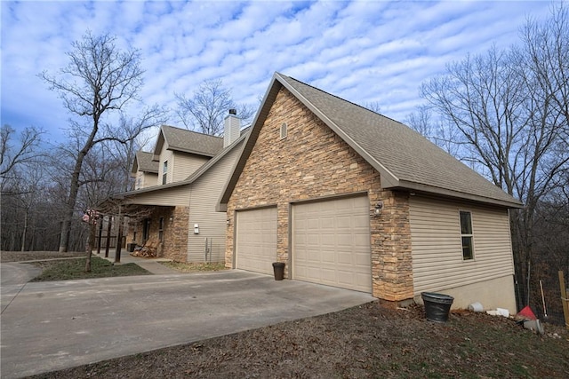 view of side of property with an attached garage, driveway, stone siding, roof with shingles, and a chimney