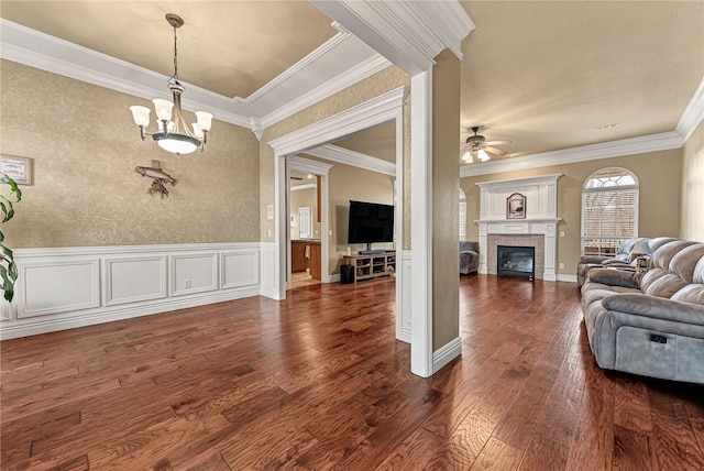 living room with dark wood-style floors, a wainscoted wall, crown molding, a tiled fireplace, and ceiling fan with notable chandelier