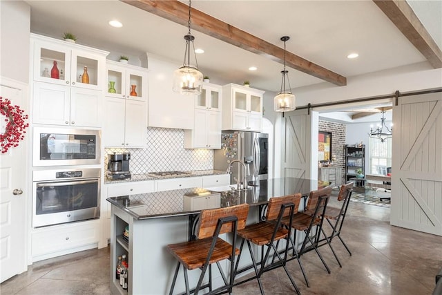 kitchen with beam ceiling, tasteful backsplash, a barn door, appliances with stainless steel finishes, and a kitchen island with sink