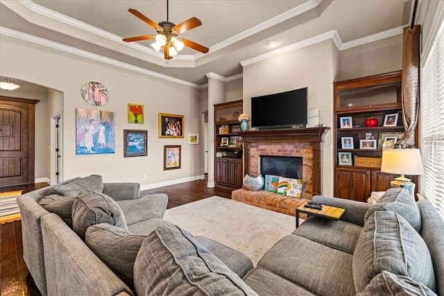 living area with dark wood-style flooring, a raised ceiling, ornamental molding, a brick fireplace, and baseboards