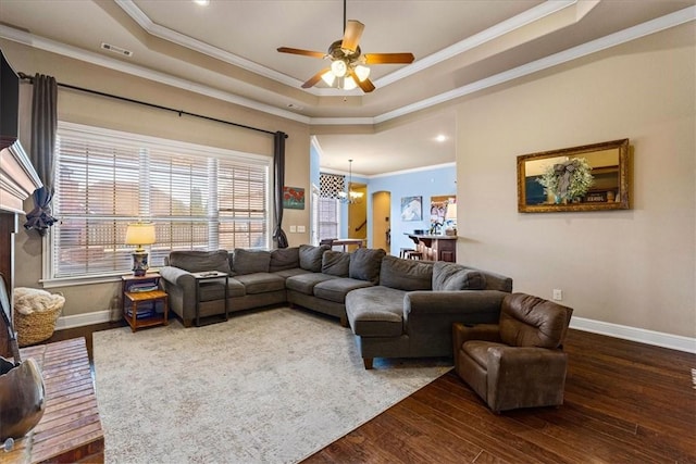 living area featuring a tray ceiling, ornamental molding, wood finished floors, baseboards, and ceiling fan with notable chandelier