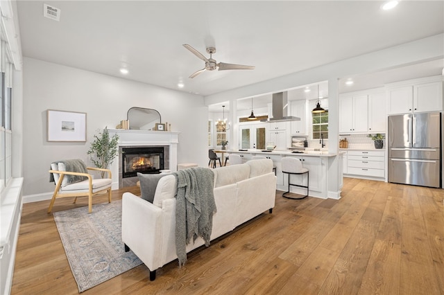 living area featuring visible vents, a glass covered fireplace, ceiling fan with notable chandelier, light wood-style floors, and recessed lighting