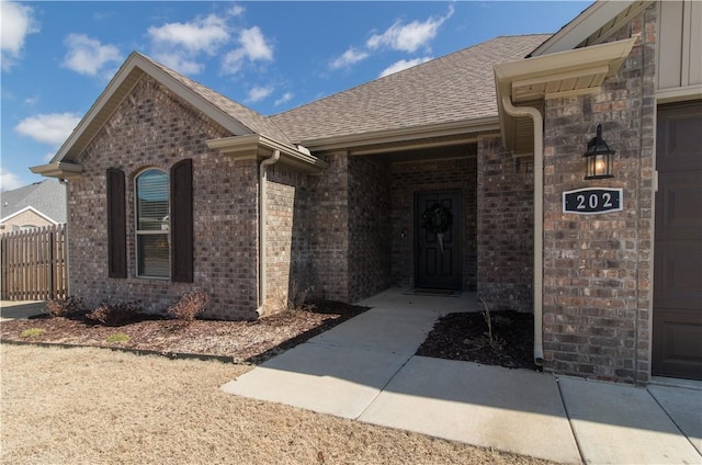 view of exterior entry featuring brick siding, a shingled roof, and fence