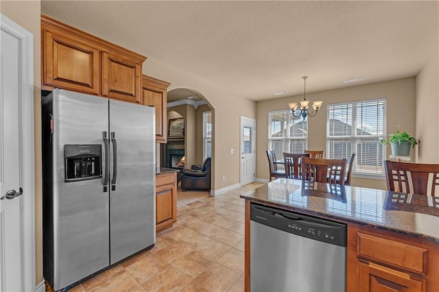 kitchen with arched walkways, brown cabinets, appliances with stainless steel finishes, dark stone countertops, and a warm lit fireplace