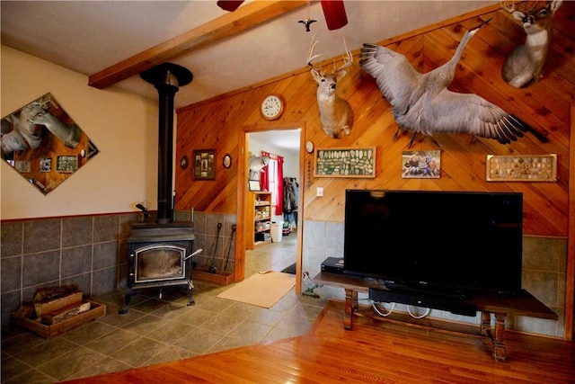 tiled living area featuring a wood stove, wood walls, a ceiling fan, and beam ceiling