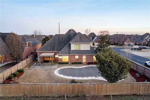 back of house at dusk with driveway, a fenced front yard, a shingled roof, brick siding, and a patio area