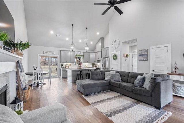 living area featuring high vaulted ceiling, a ceiling fan, a fireplace with raised hearth, and wood finished floors