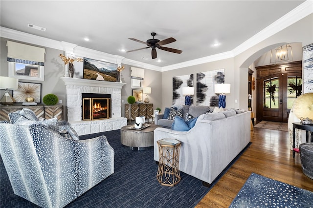 living area featuring arched walkways, a stone fireplace, wood finished floors, visible vents, and ornamental molding