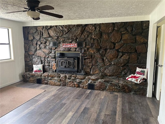 unfurnished living room with a textured ceiling, ceiling fan, wood finished floors, and a wood stove