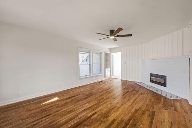 unfurnished living room with built in features, a ceiling fan, wood finished floors, a tile fireplace, and baseboards