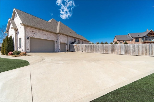 view of side of property with fence, driveway, a shingled roof, a garage, and brick siding