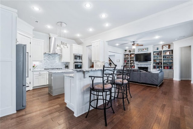 kitchen with a breakfast bar, appliances with stainless steel finishes, wall chimney exhaust hood, a fireplace, and ceiling fan