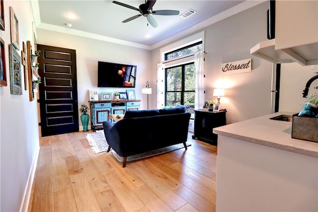 living room with light wood finished floors, visible vents, baseboards, ceiling fan, and ornamental molding