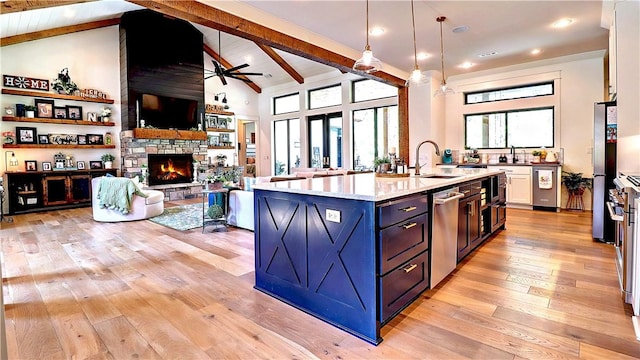 kitchen featuring beam ceiling, a ceiling fan, light wood-style floors, appliances with stainless steel finishes, and a stone fireplace