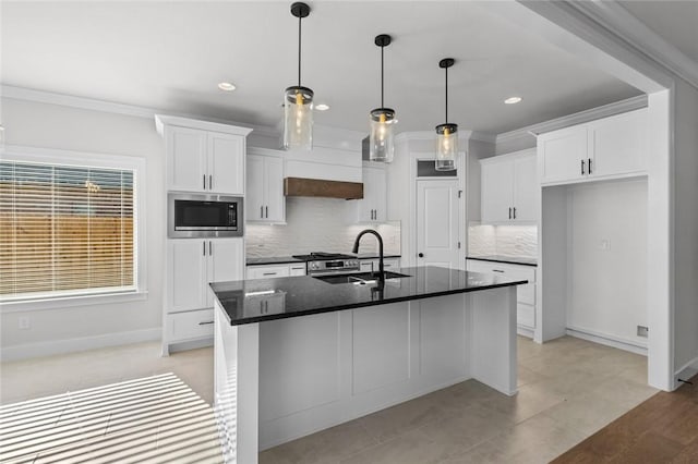 kitchen with stainless steel microwave, a center island with sink, ornamental molding, white cabinetry, and a sink