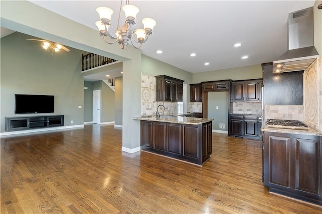 kitchen with dark brown cabinets, light stone countertops, open floor plan, wall chimney exhaust hood, and a sink