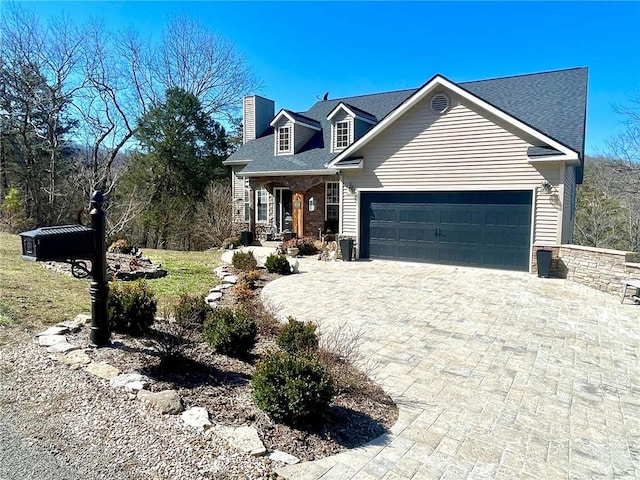 cape cod house with decorative driveway, an attached garage, a shingled roof, and a chimney