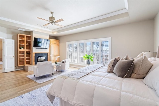 bedroom featuring ceiling fan, light wood-style flooring, a raised ceiling, and a glass covered fireplace