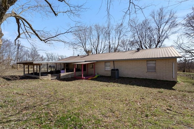 back of house with a yard, central AC, a chimney, brick siding, and metal roof