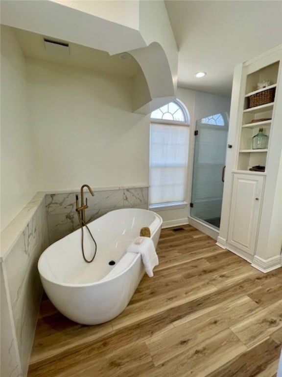 full bath featuring wood finished floors, a wainscoted wall, a freestanding tub, a shower stall, and tile walls