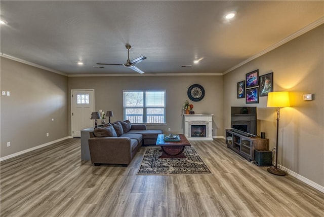 living room featuring ornamental molding, wood finished floors, a glass covered fireplace, baseboards, and ceiling fan