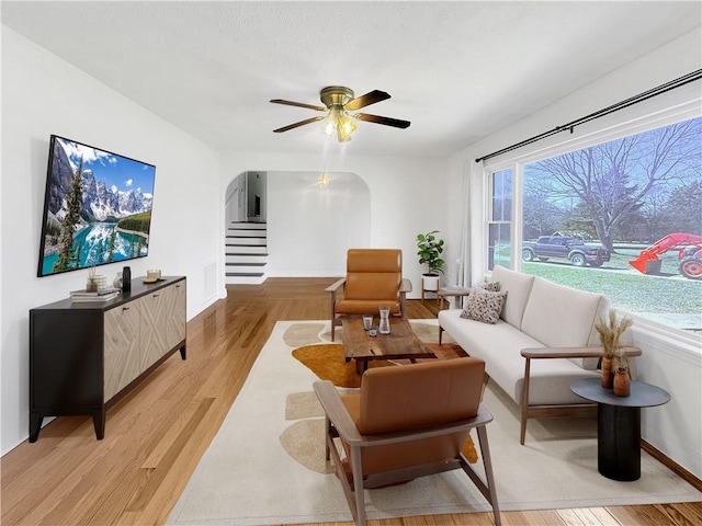 living room featuring baseboards, stairs, light wood-style floors, arched walkways, and a ceiling fan