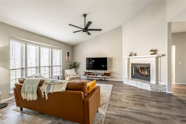 living area with high vaulted ceiling, a ceiling fan, a glass covered fireplace, wood finished floors, and baseboards