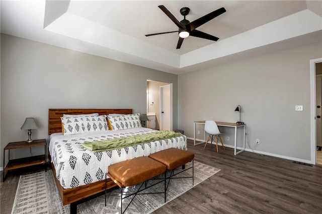 bedroom featuring wood finished floors, baseboards, a tray ceiling, ceiling fan, and ensuite bathroom