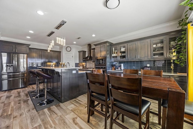 kitchen featuring a breakfast bar, light countertops, glass insert cabinets, appliances with stainless steel finishes, and wall chimney range hood