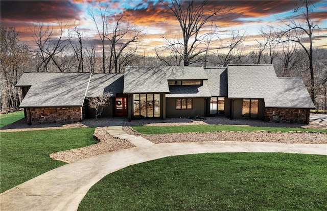 mid-century home featuring a yard, stone siding, and roof with shingles