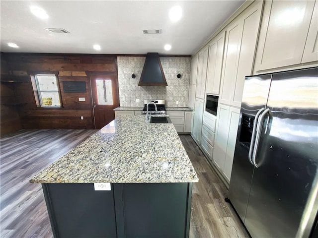 kitchen with light stone counters, custom exhaust hood, stainless steel appliances, dark wood-type flooring, and wood walls