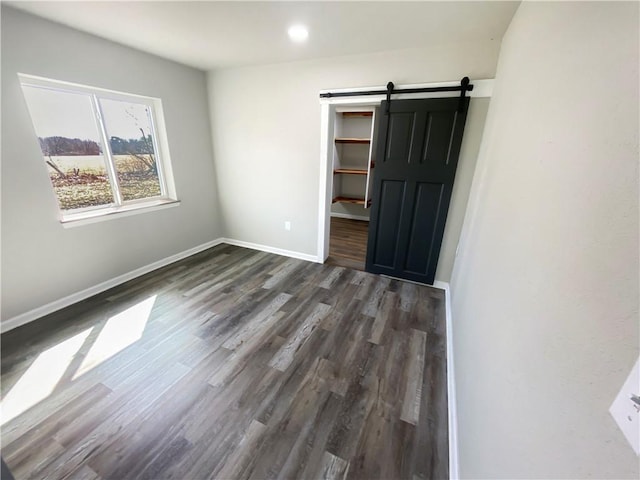 unfurnished bedroom with a walk in closet, baseboards, a barn door, recessed lighting, and dark wood-style flooring