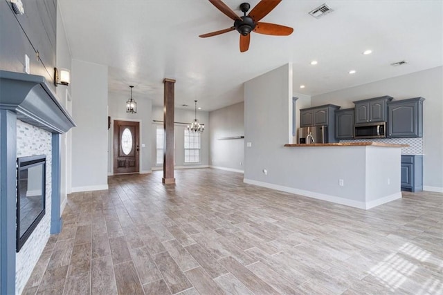 unfurnished living room featuring visible vents, decorative columns, a fireplace, light wood-style floors, and ceiling fan with notable chandelier