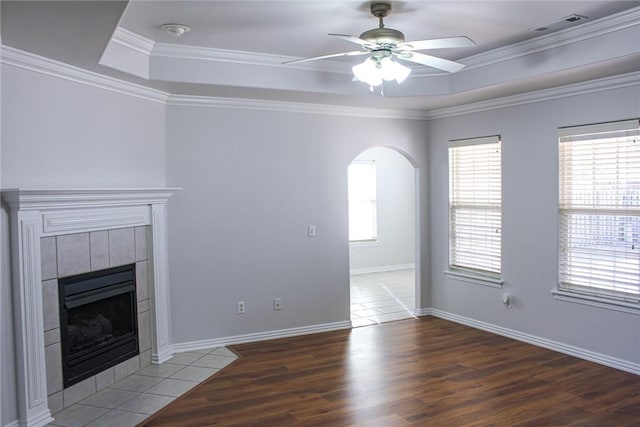 unfurnished living room with wood finished floors, arched walkways, crown molding, a tiled fireplace, and a raised ceiling