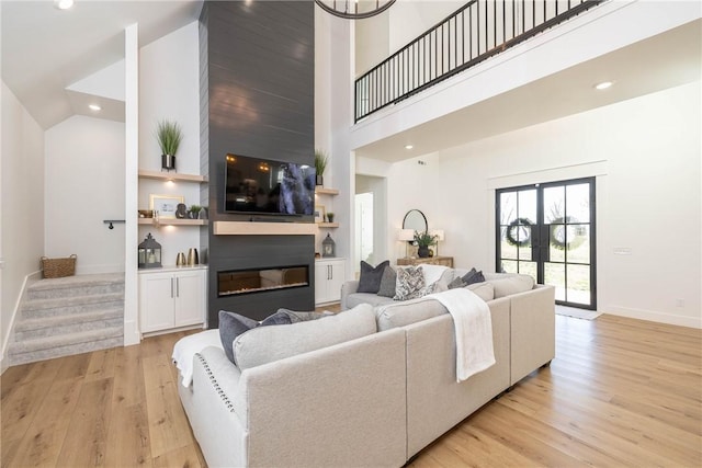 living area with a glass covered fireplace, stairway, light wood-style floors, baseboards, and a towering ceiling