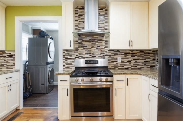 kitchen featuring tasteful backsplash, wall chimney range hood, light wood-style flooring, appliances with stainless steel finishes, and stacked washing maching and dryer