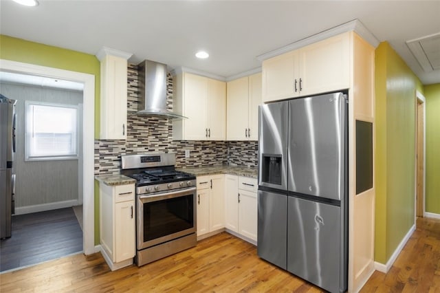 kitchen with light wood-type flooring, light stone counters, backsplash, appliances with stainless steel finishes, and wall chimney range hood