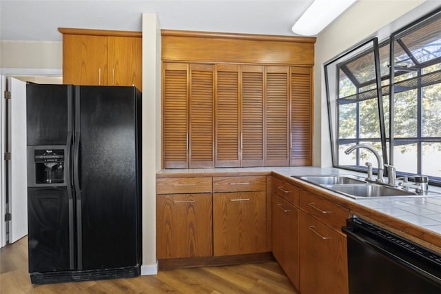 kitchen featuring light wood-style flooring, a sink, black appliances, tile counters, and brown cabinets